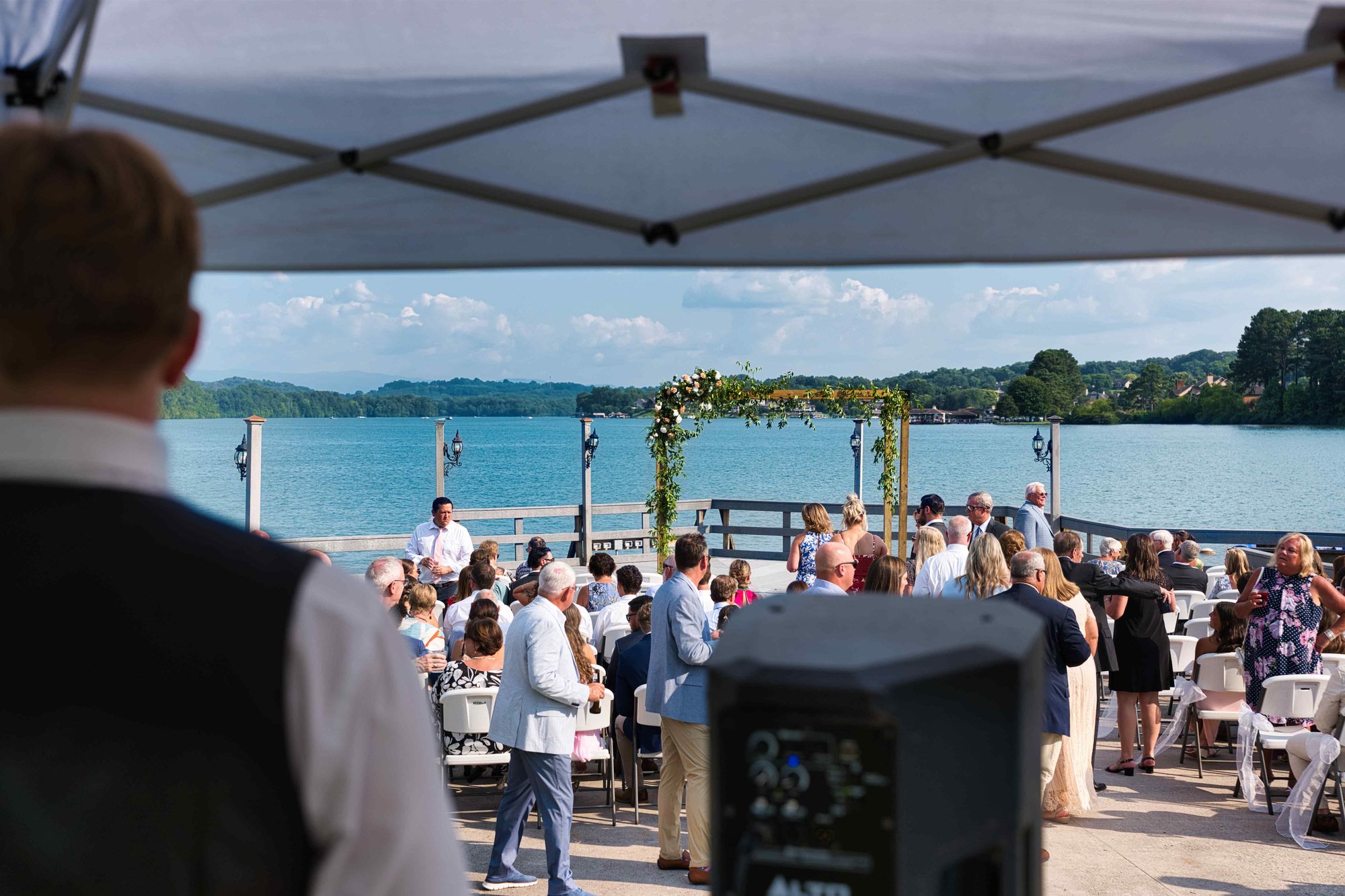 A lakeside wedding ceremony seen past a speaker on a stand, the ceremony sound already in position