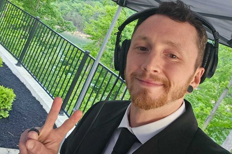 Coty Tate in a black suit and tie with headphones on, smiling under an event tent with trees and a lake behind him