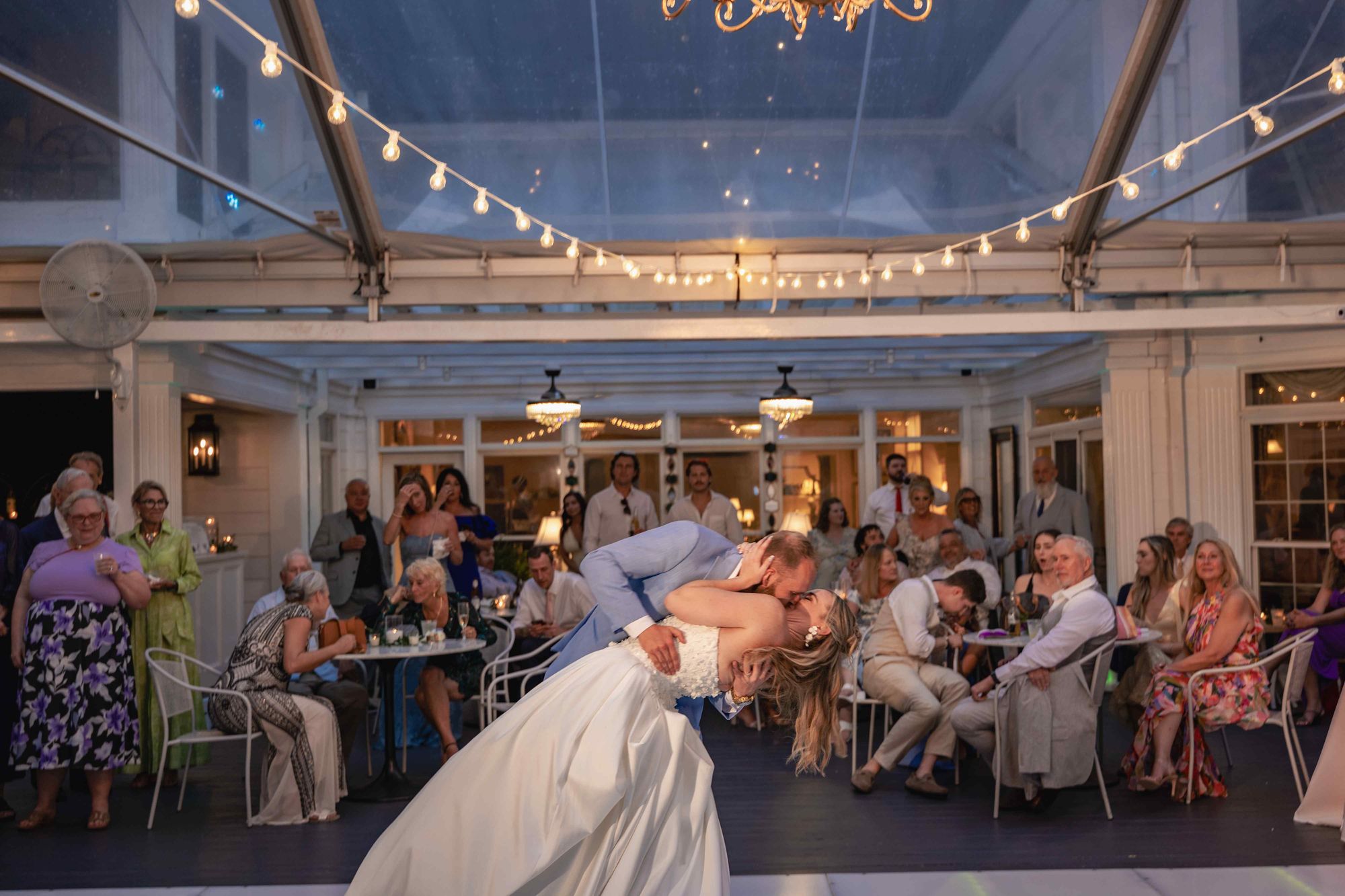 A couple kissing in a dip beneath strings of bistro lighting, guests seated around the floor
