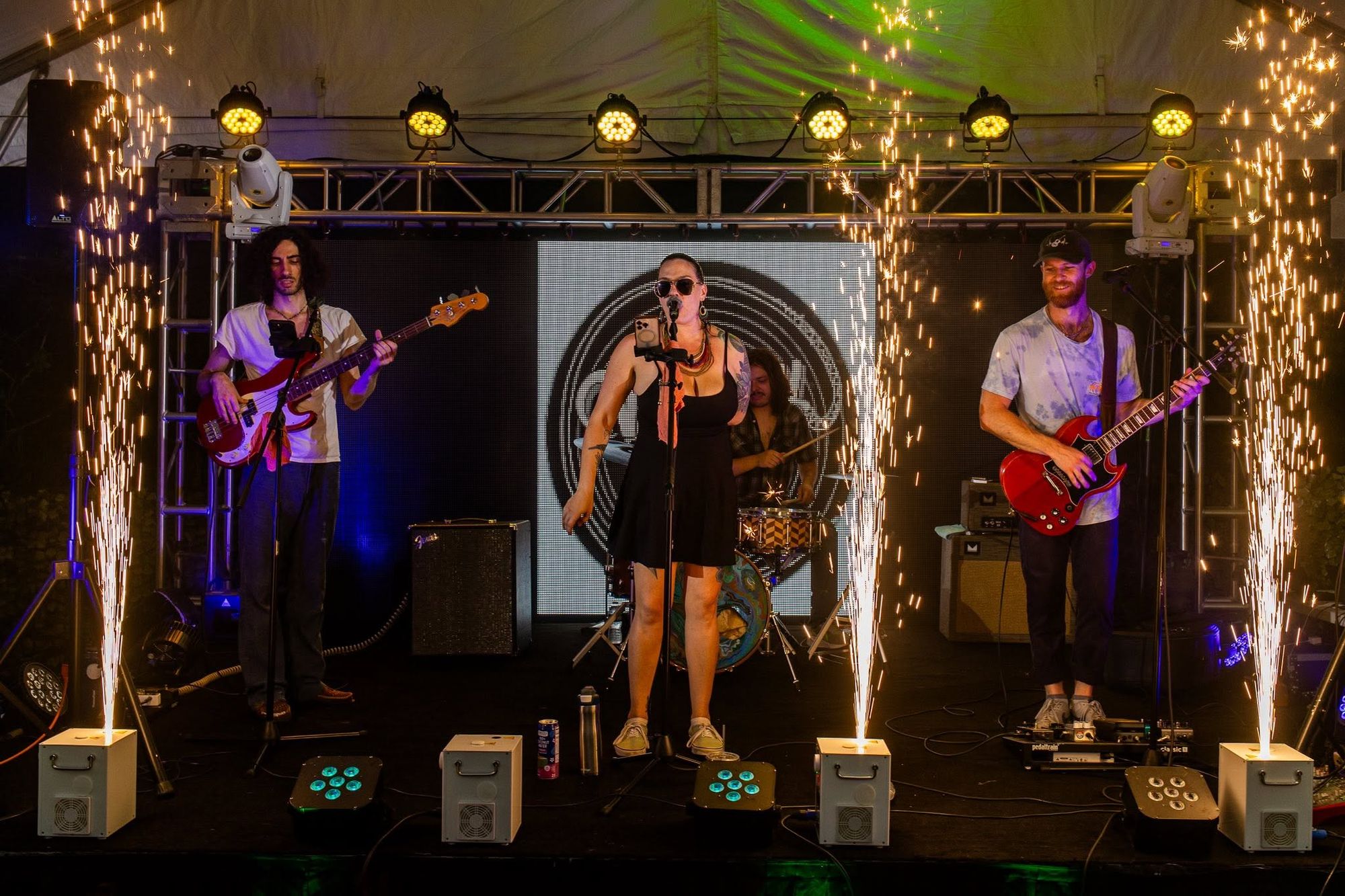 Six cold spark machines firing fountains of sparks along the front of a tented stage while a four-piece band plays