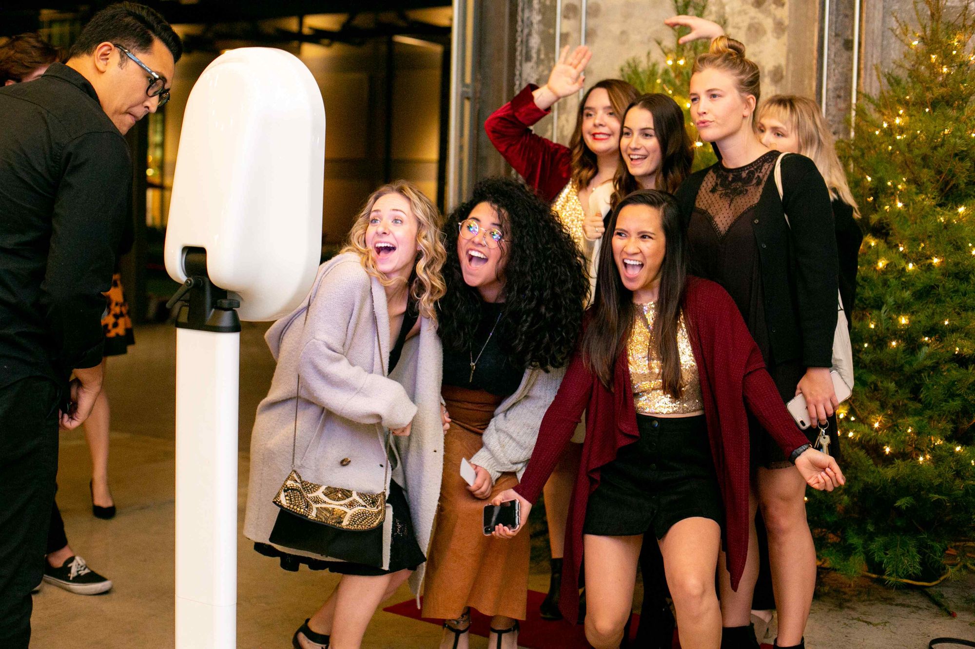 Six guests crowded in front of an open-air photo booth mid-laugh, arms up, with the party and a lit tree behind them