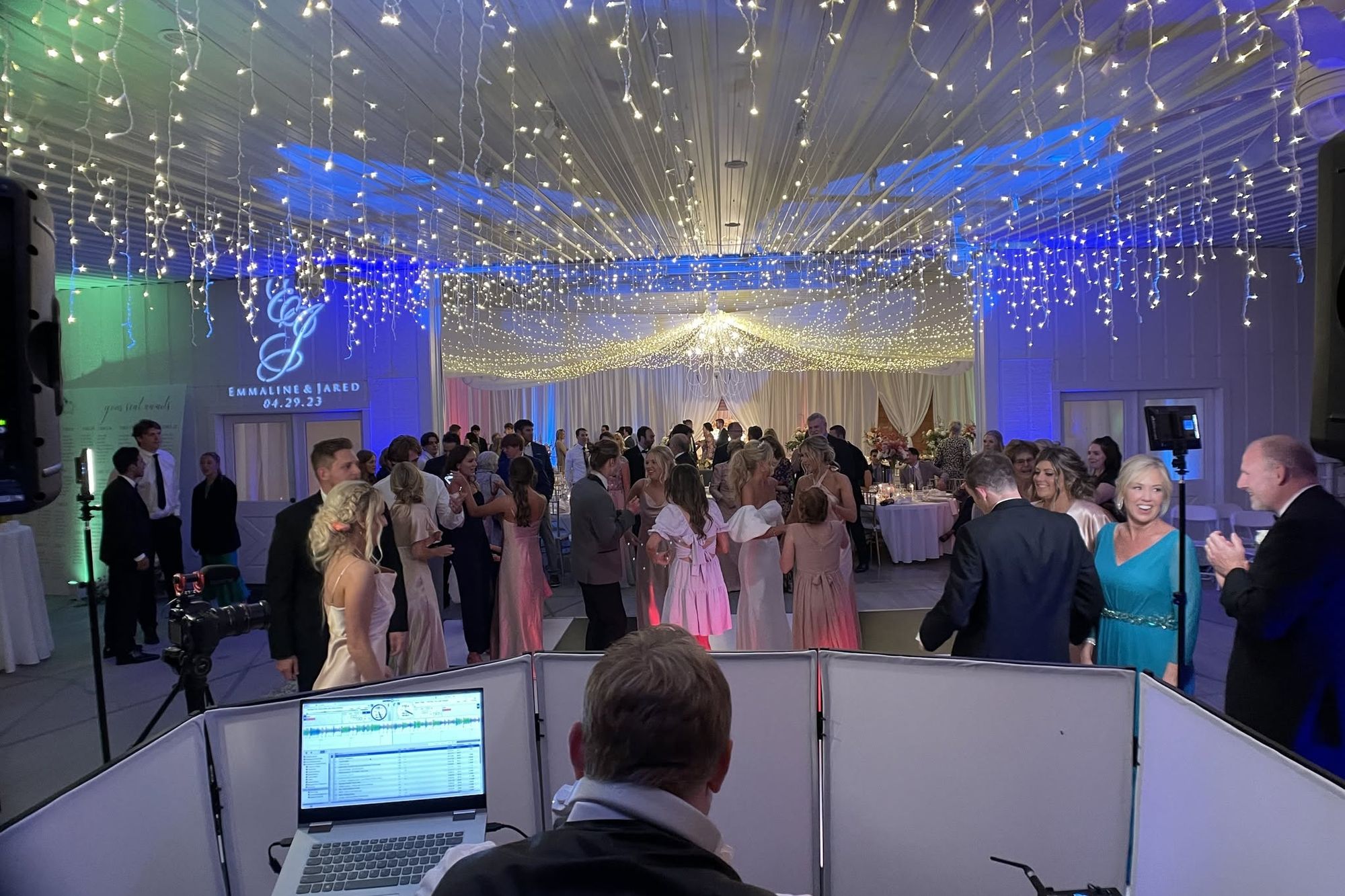 A projected monogram reading E and J with the couple’s names and date, lit on a wall beside a reception floor under strings of icicle lighting