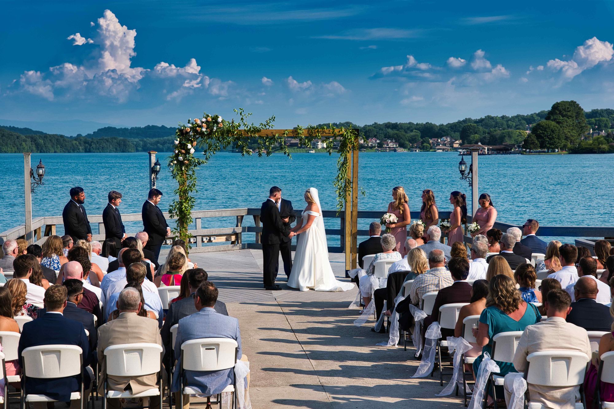 A wedding ceremony on a pier, guests seated and the lake open behind the arch