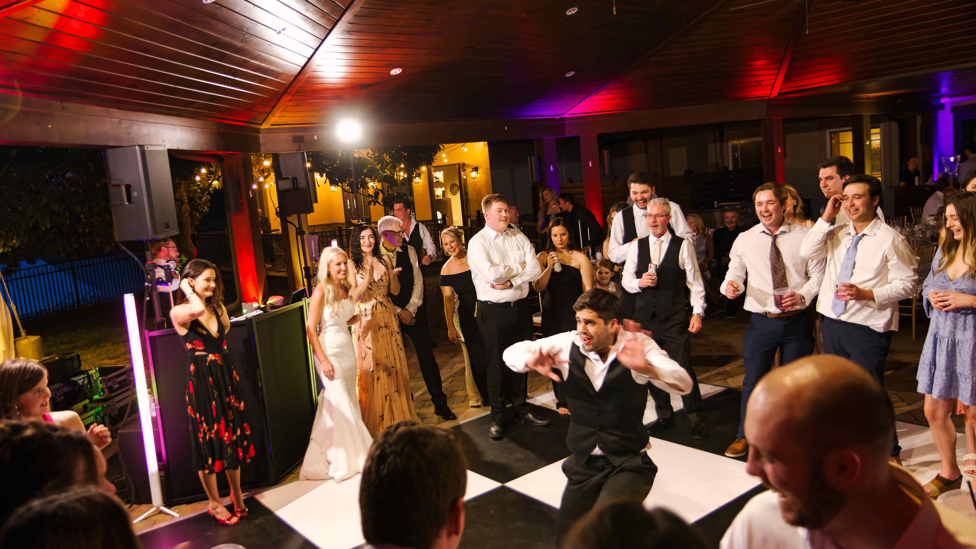 A guest mid-dance on a checkered floor at a wedding reception, the room lit in purple and amber and the crowd laughing around him
