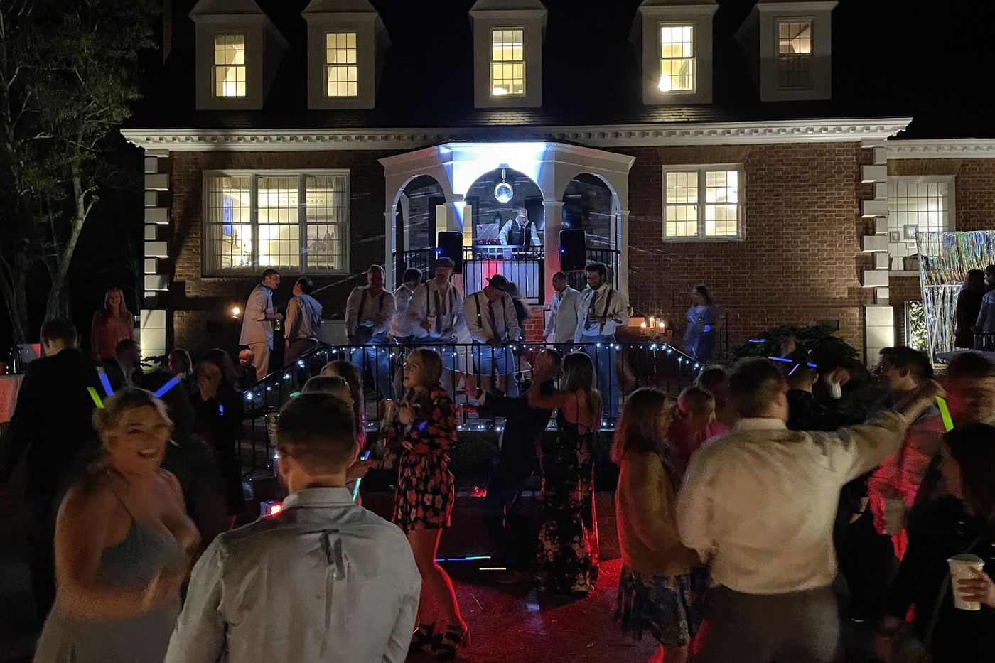 A party in the drive of a private house at night, the DJ set up on the porch under a mirror ball and the crowd dancing below with light sticks
