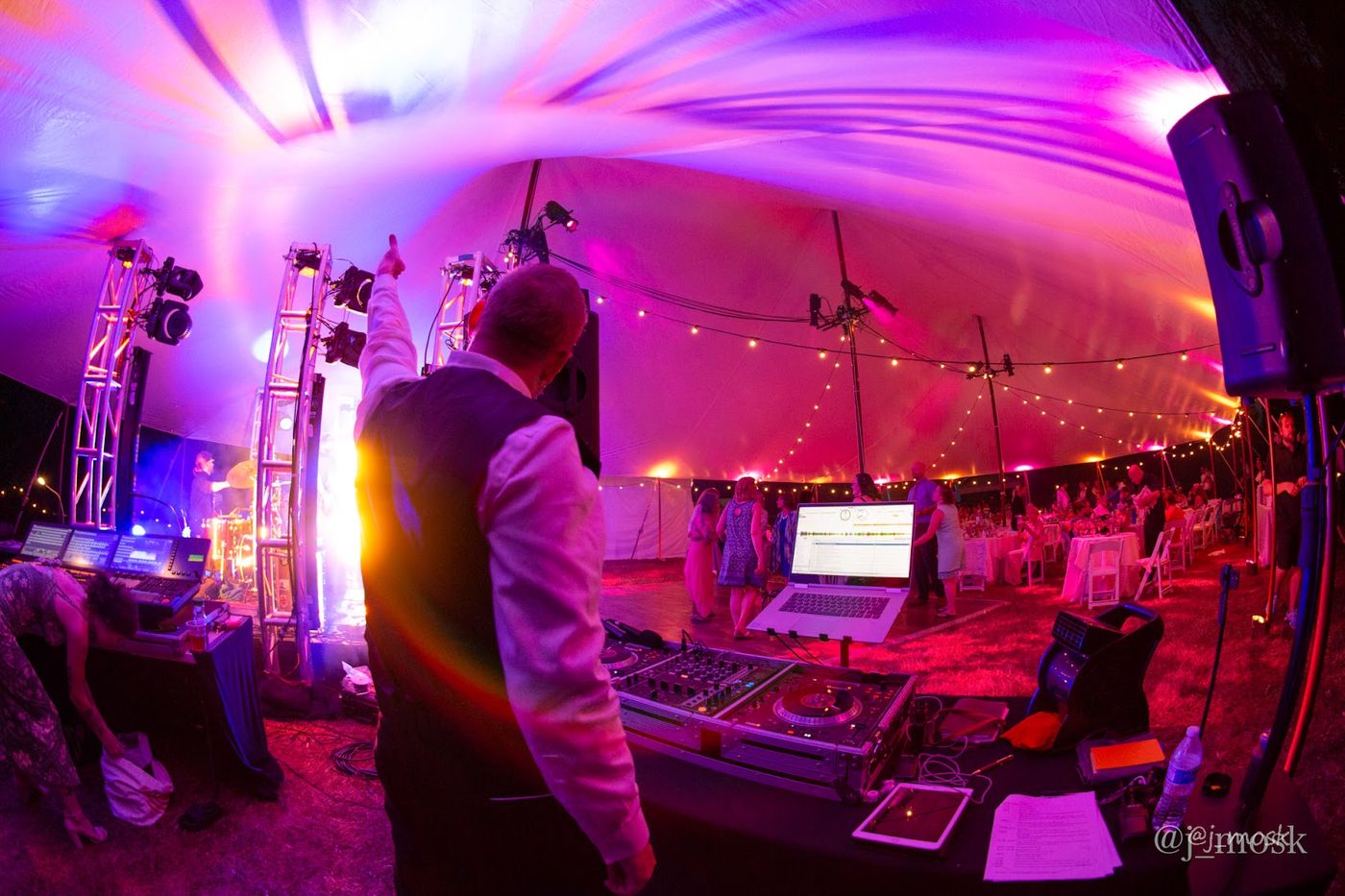 Rob at the booth under a tent lit magenta, one arm raised to the room, with the lighting truss, the mixer and the run of show laid out in front of him