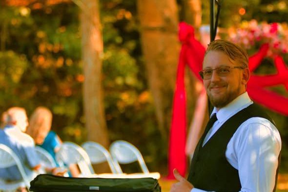Rob Kermidas in shirt, tie and waistcoat beside the ceremony sound at an outdoor wedding, the chairs set and the arch dressed behind him