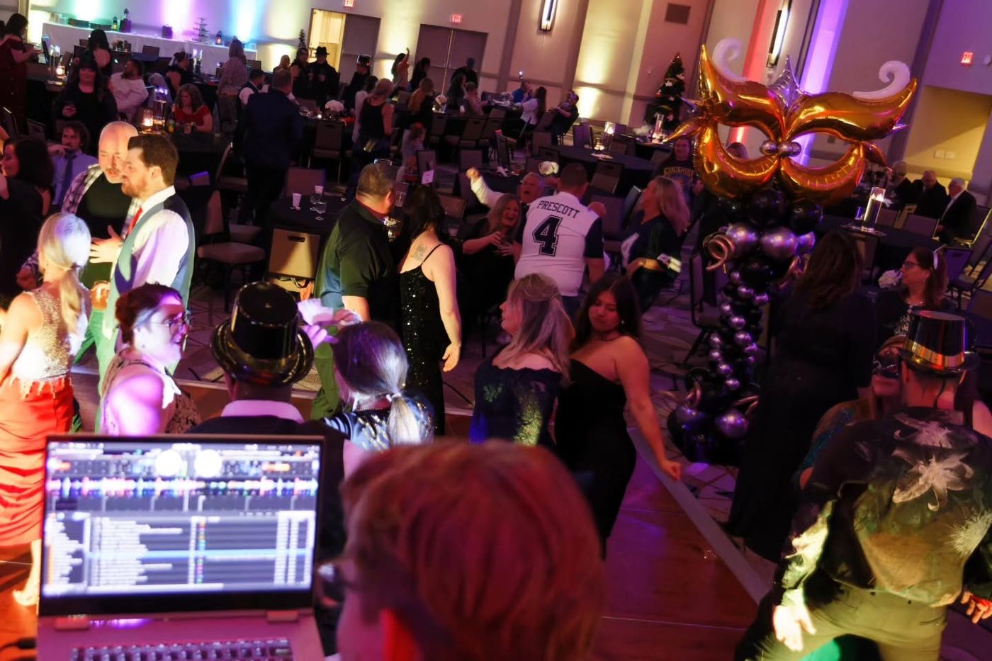 A masquerade ball in a hotel ballroom, the walls uplit and the floor filling, seen from behind the DJ booth