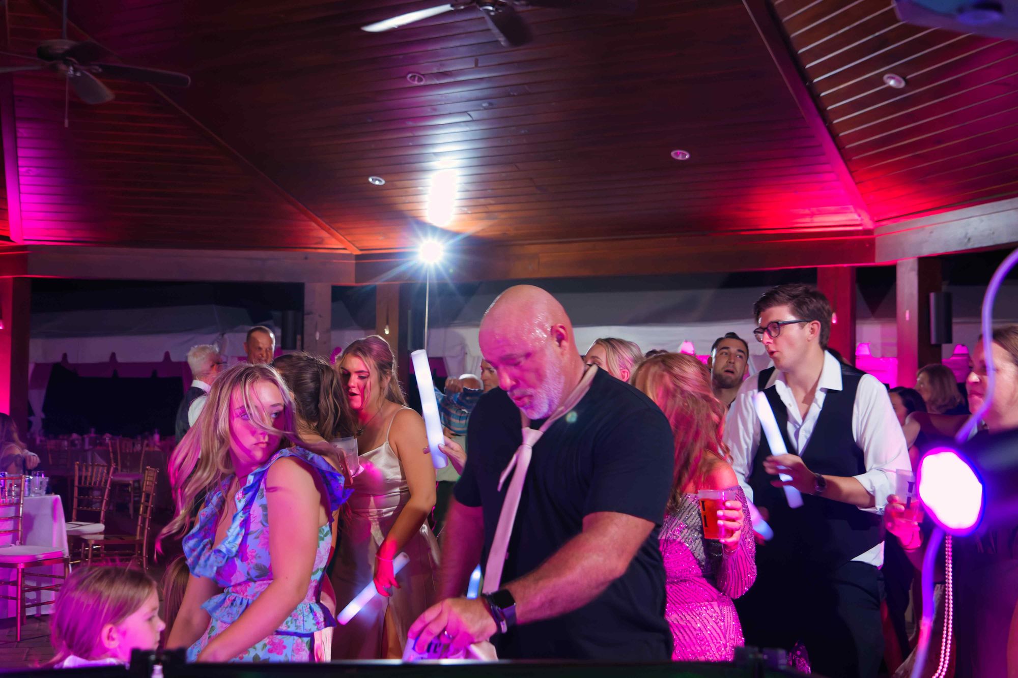 Guests of every age dancing together under magenta uplighting, light sticks in the air