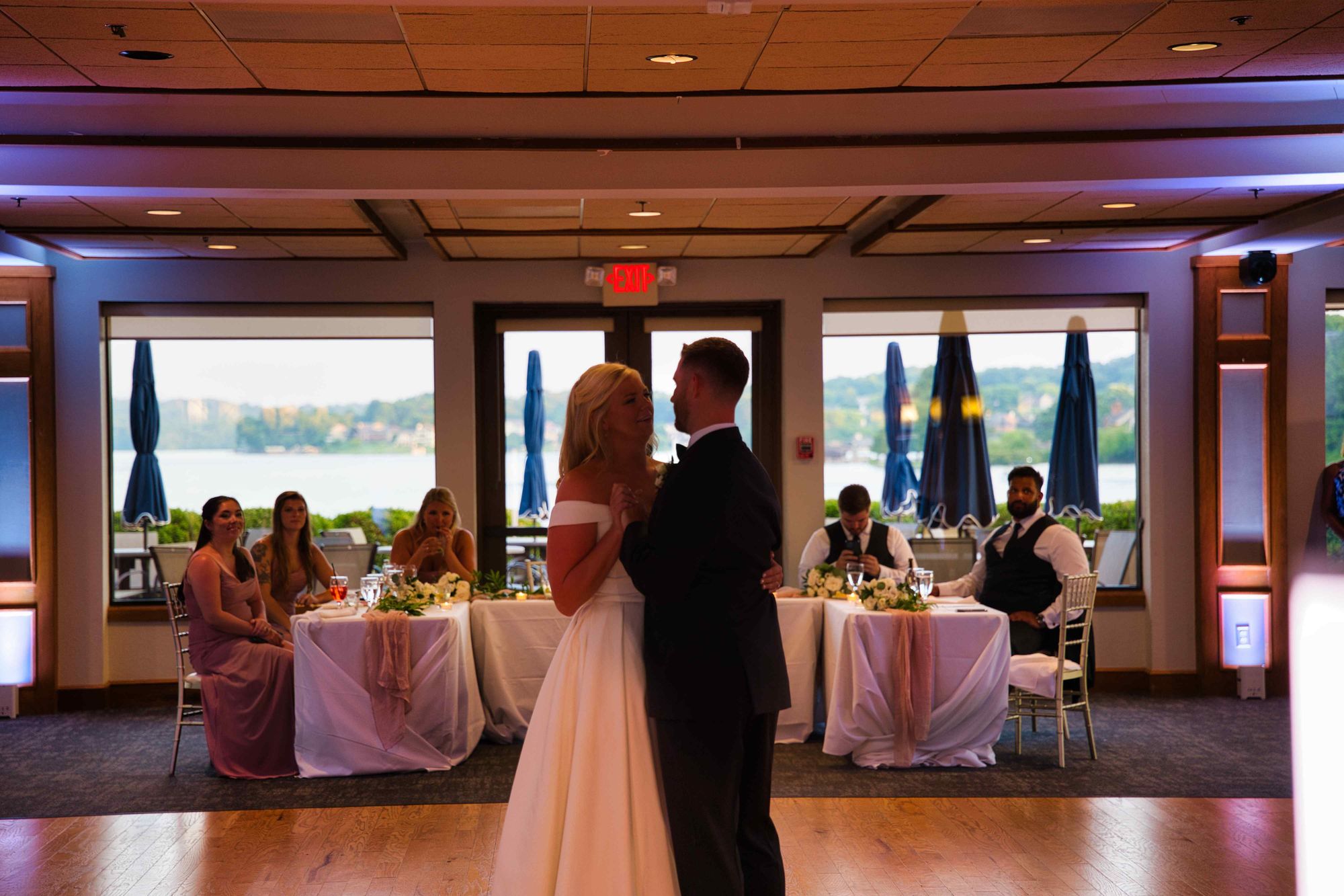 A couple dancing their first dance on a wood floor, with the lake visible through the windows behind them
