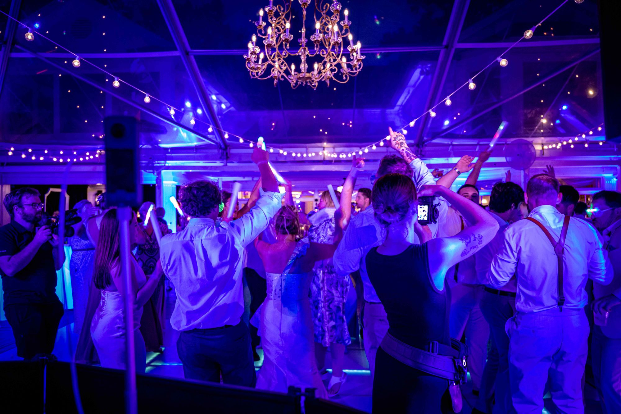 A packed dance floor under a chandelier and string lighting, the room washed in deep blue